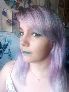 [Justine CM] Photo de moi avec les cheveux couleur lilas pâle. Je porte un rouge à lèvres turquoise, du crayon à paupières noir et du fard à paupières vert émeraude. Je porte une robe blanche à fleurs roses, dont des roses, et un pendentif topaze bleu clair en forme de cœur.