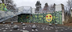 [Justine CM] Streetart sur Essert, sur un parc de jeux. Pris en deux photos. Photo 2 : Fond vert, sur lequel il est écrit en majuscules remplies de prénoms "Émotion", avec le dernier "O" en forme de smiley jaune qui a des yeux en forme de cœur et qui sourit.