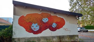 [Justine CM] Streetart à Dardel, Belfort. Trois têtes blanches, aux traits asiatiques, sont dessinées, et sont dans un nuage orangé avec des aspects ronds sous forme d'arabesques. Art par Clémentine Martinez.