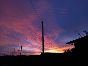 [Justine CM] Photo d'un ciel à l'aube. Le bleu doux laisse place à des nuages roses et au fond, le soleil flamboie d'une aura orangée. Poteaux électriques.