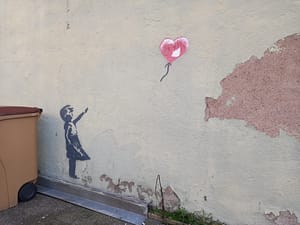 [Justine CM] Streetart dans une rue parallèle à la rue Jean Jaurès à Belfort. Une petite fille en robe est dessinée en noir, et a laissé échapper un ballon rouge en forme de cœur.