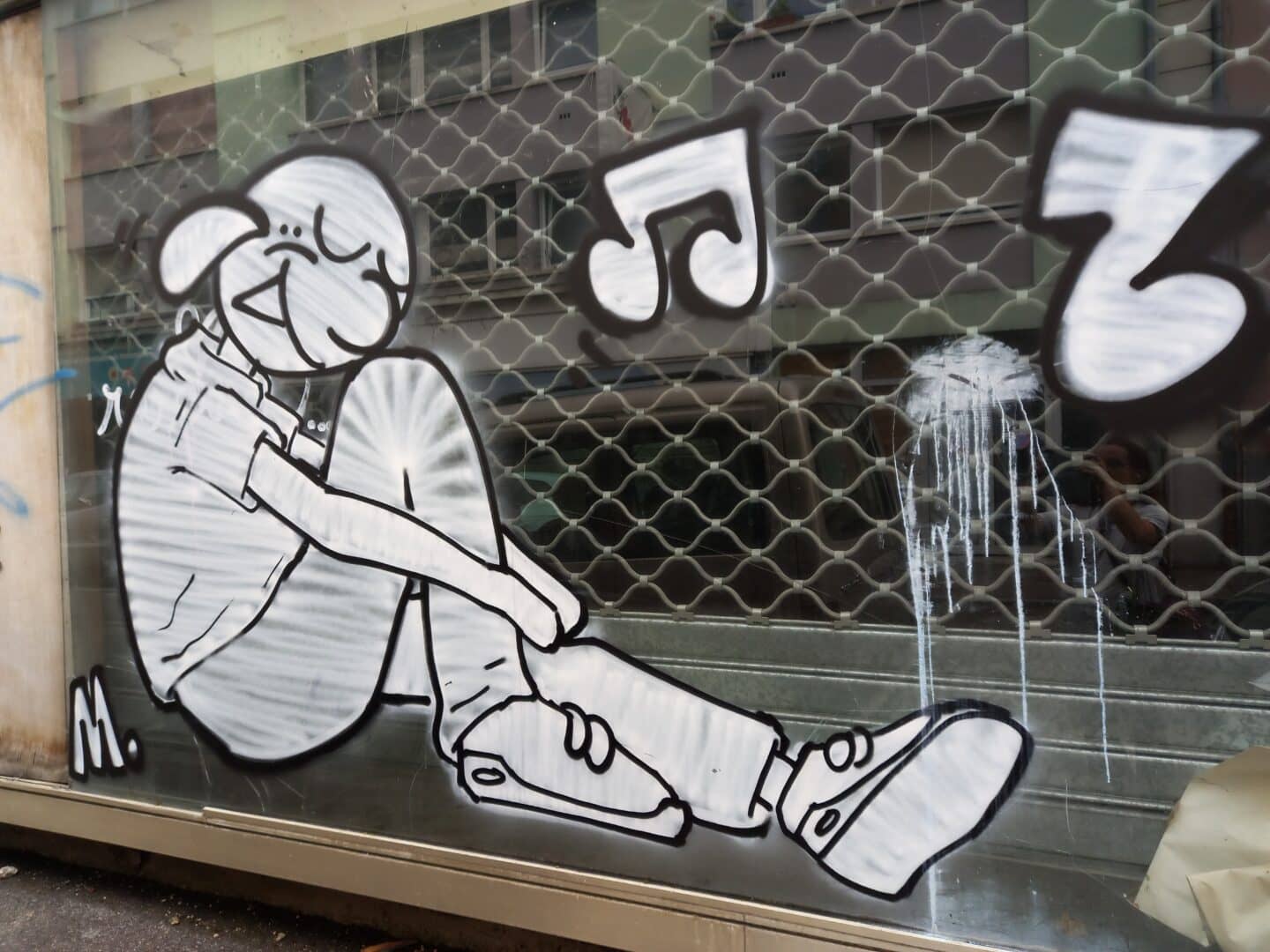 [Justine CM] Belfort, rue parallèle à la rue piétonne et la gare. Photo prise en août 2022. Personnage dessiné sur la vitre d'un ancien magasin, assis, un genou relevé, souriant, tout en blanc. Note de musiques autour.